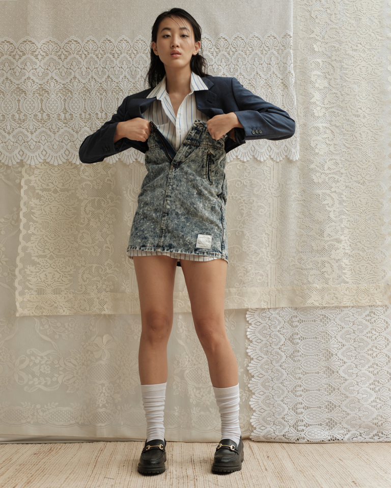 Model wearing a cropped navy blazer from Now Thenn styled over a striped shirt and acid-wash denim dress, paired with white socks and black loafers, photographed against a lace backdrop.