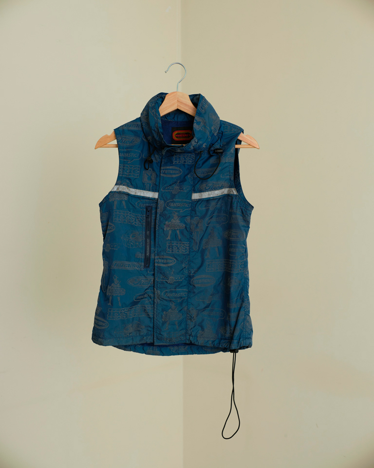 Blue reflective utility vest with glow-in-the-dark detailing, graphic print, high-neck collar, zip front, and drawcord hem – worn layered for a streetwear look.