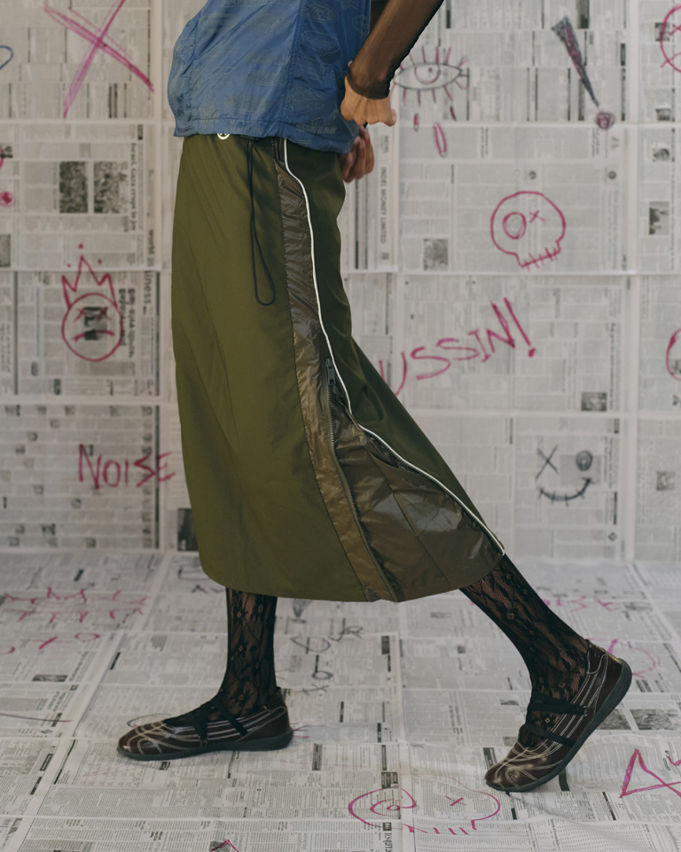 Olive utility midi skirt with side zip panels, reflective piping, elastic waist, and relaxed fit, styled with layered streetwear looks.