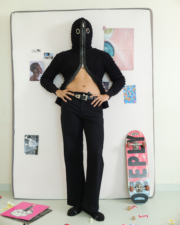 Black textured zip hood streetwear jacket with metal eye rivet detail on hood and full front zipper, unisex techno-inspired statement outerwear styled with wide-leg trousers.